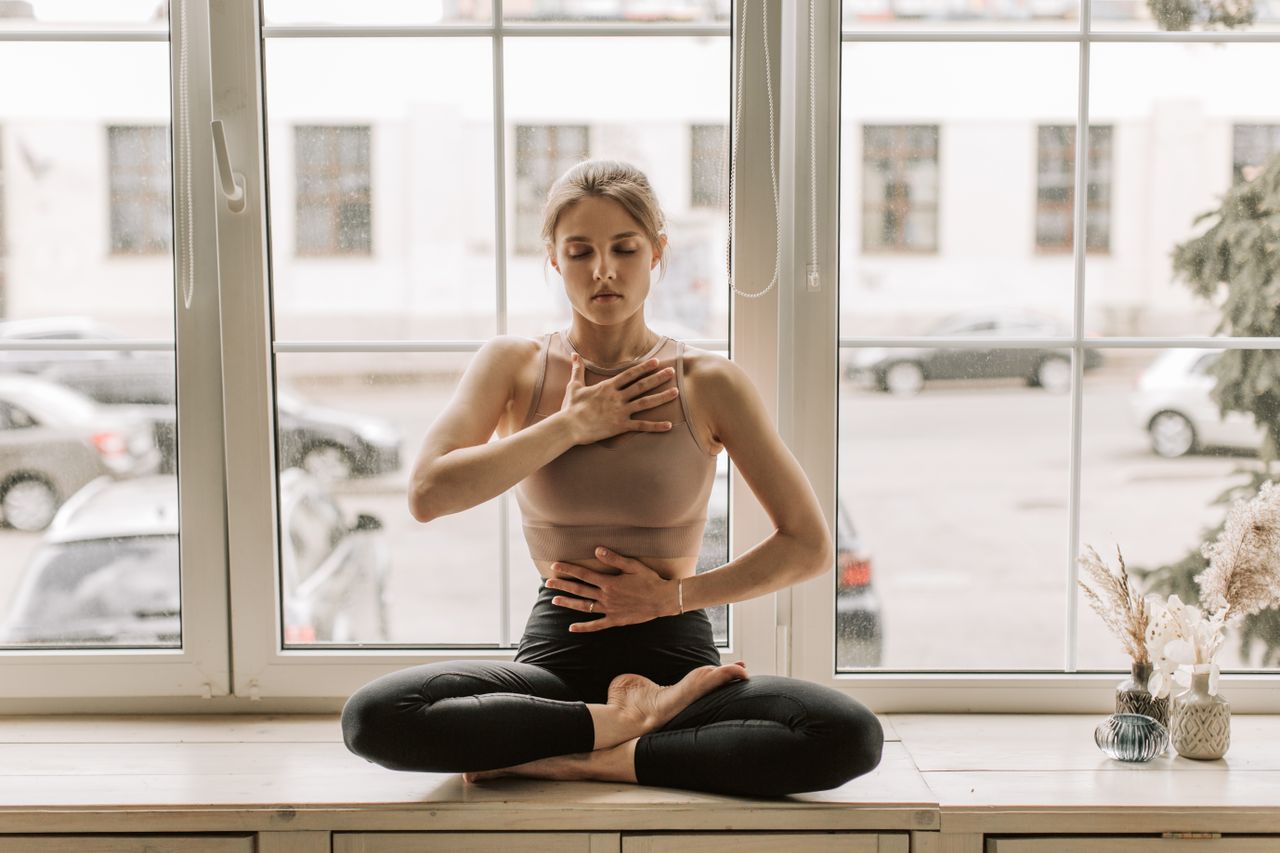 A woman practicing meditation in a serene yoga pose by a large window.